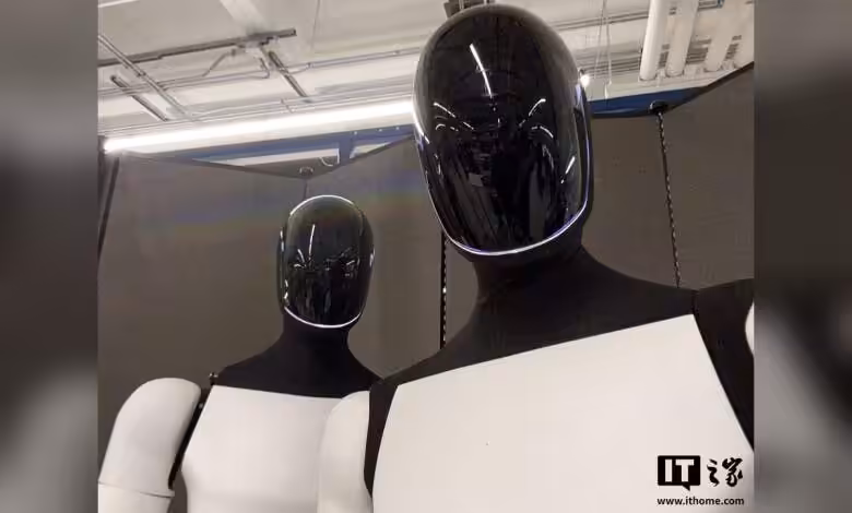 Tesla Optimus reculé en R&D : Musk admet aucun robot utile en production 1 Deux robots humanoïdes Tesla Optimus aux visages noirs brillants et torses blancs dans un environnement industriel.