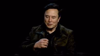 Homme en veste sombre tenant un microphone sur un fond noir.