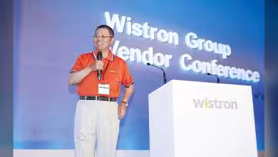 Conférence Wistron, présentateur en polo rouge sur scène, logo Wistron en arrière-plan.