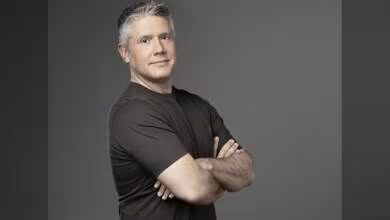 Portrait d'un homme aux cheveux gris, en t-shirt foncé, bras croisés, fond neutre.