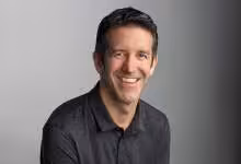 Homme souriant en chemise grise sur fond uni, cheveux courts foncés.