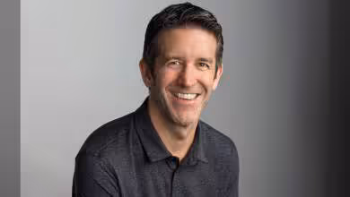 Homme souriant en chemise grise sur fond uni, cheveux courts foncés.