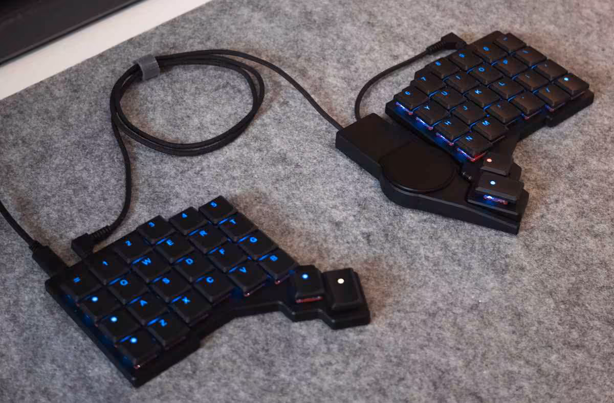 Deux claviers ergonomiques ZSA connectés, touches rétroéclairées bleues, posés sur tapis gris.