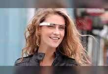 Femme portant lunettes connectées Google Glass, cheveux blonds, veste noire, en extérieur.