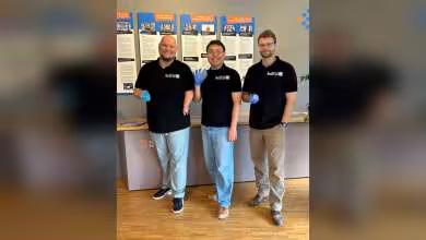 Trois hommes devant des posters NexiGO, portant t-shirts noirs et gants bleus, présentation de laboratoire réelle