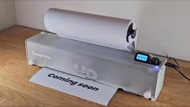 Imprimante OpenPrinter transparente affichant une feuille “coming soon” et un écran de contrôle numérique.
