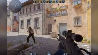 Capture d’écran d’un jeu de tir à la première personne, fusil sniper en main, interface HUD visible, village méditerranéen.