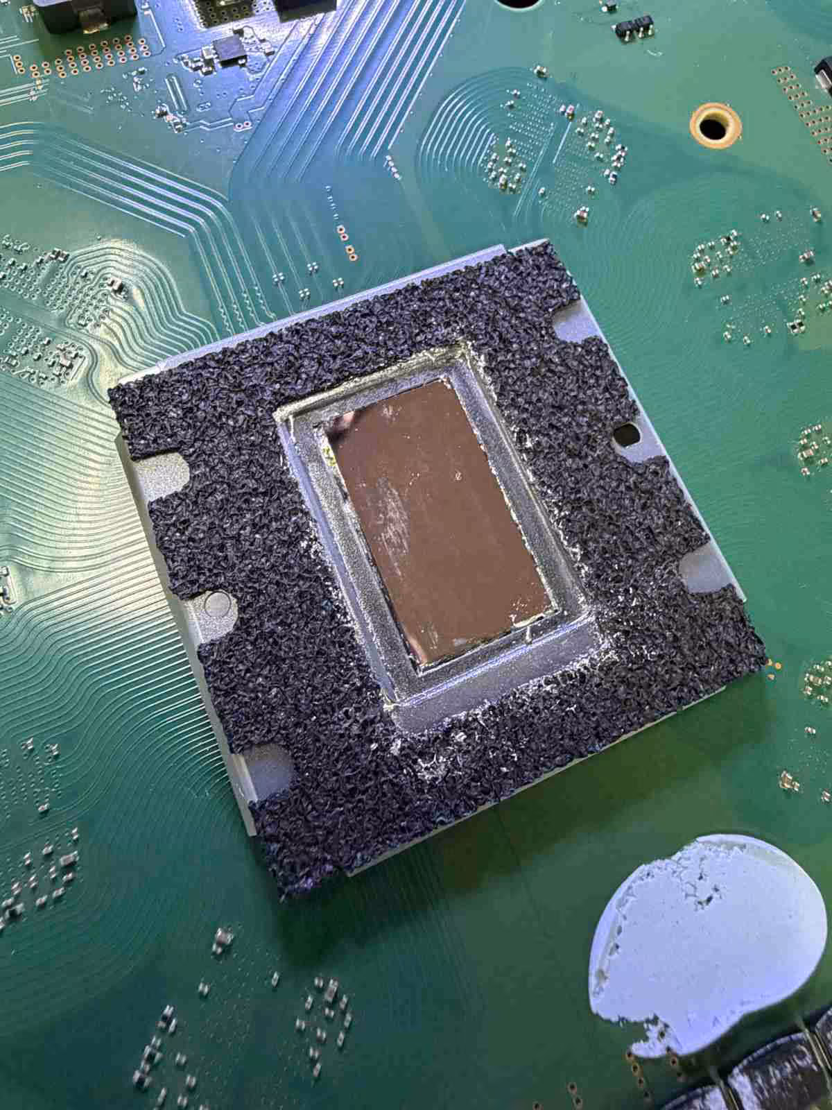 PS5 : une réparation montre une corrosion du dissipateur sous le métal liquide 2 Vue rapprochée d’un radiateur PS5 endommagé sur une carte verte, circuits visibles, éclairage naturel