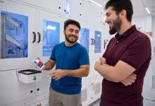 Deux employés souriants devant des machines automatisées, l’un tenant un badge ou smartphone Apple.