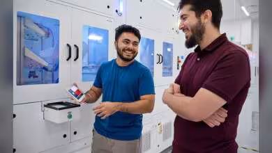 Deux employés souriants devant des machines automatisées, l’un tenant un badge ou smartphone Apple.