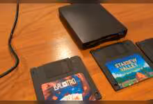 Lecteur de disquette externe avec disquettes de jeux Stardew Valley et Baldur's Gate sur bureau en bois.