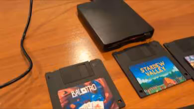 Lecteur de disquette externe avec disquettes de jeux Stardew Valley et Baldur's Gate sur bureau en bois.