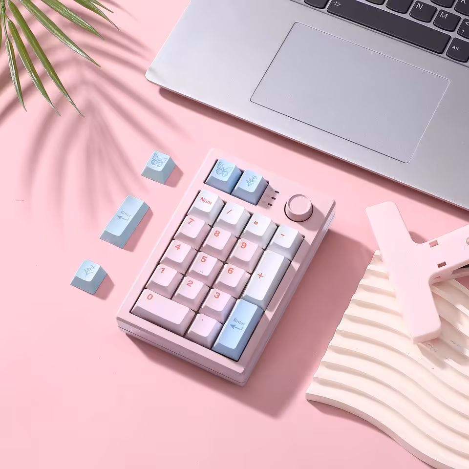 Yunzii AL21 : un pavé numérique aluminium sans fil à 49,99 dollars pour claviers compacts 15 Yunzii AL21 rose posé sur bureau pastel avec ordinateur portable, keycaps de rechange et plantes