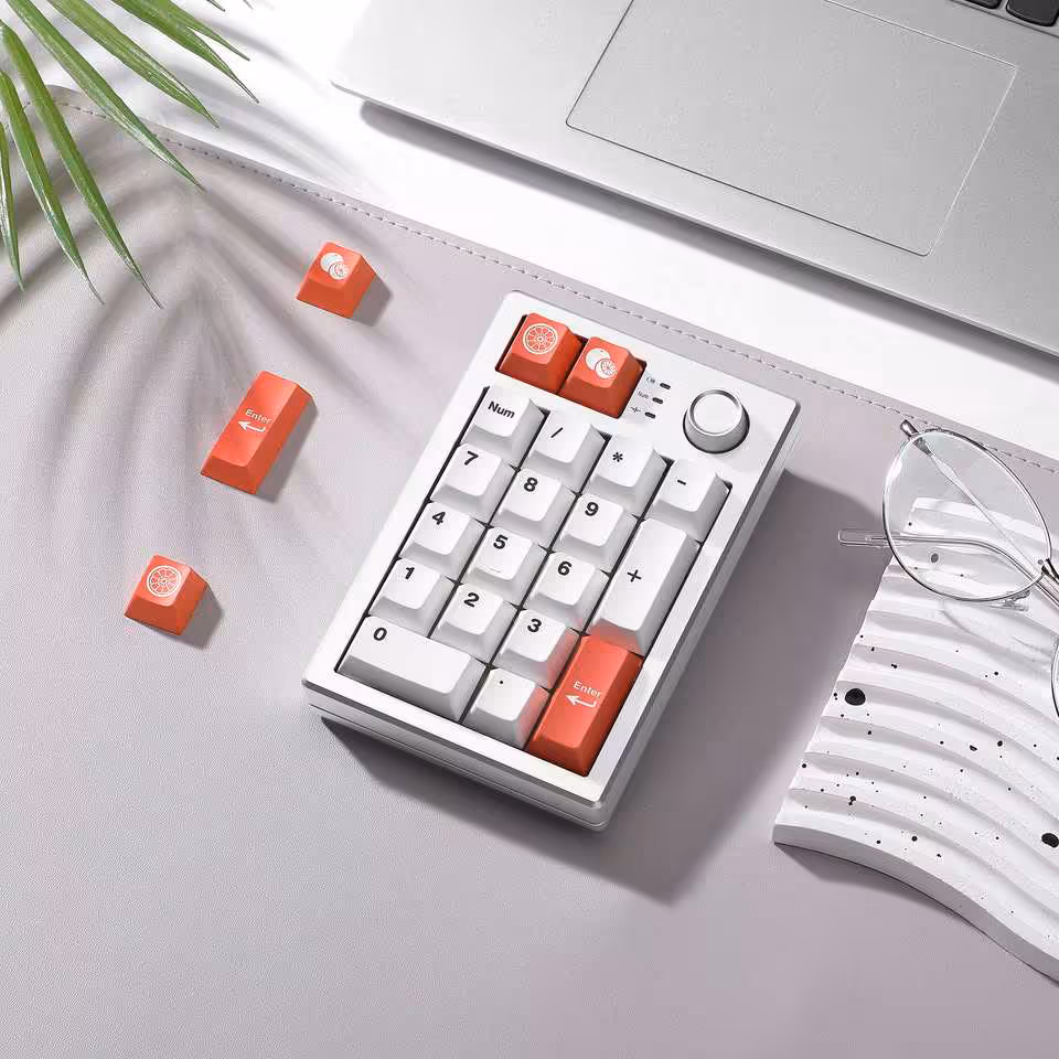 Yunzii AL21 : un pavé numérique aluminium sans fil à 49,99 dollars pour claviers compacts 16 Yunzii AL21 blanc avec keycaps orange, posé près d’un ordinateur portable et de lunettes
