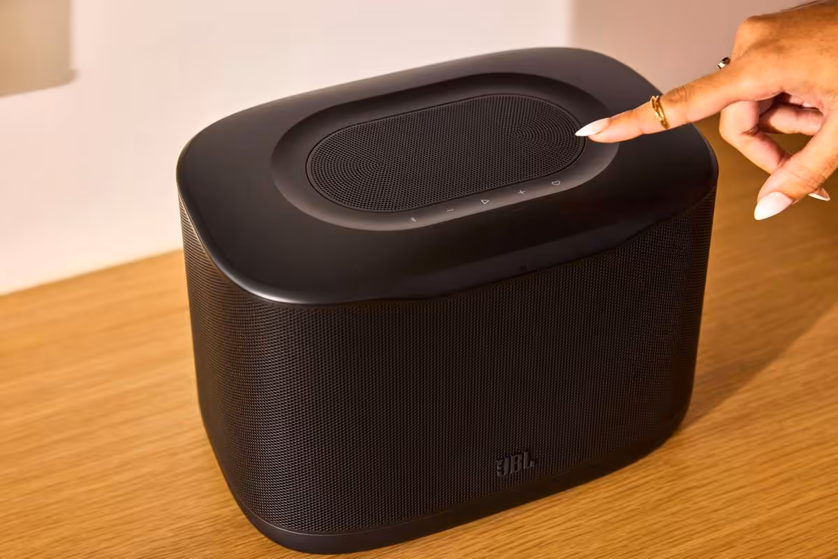 Main touchant le dessus d’une enceinte JBL Cove noire posée sur une table en bois.