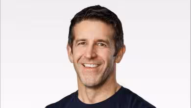 Portrait de John Ternus, dirigeant Apple, souriant sur fond blanc, t-shirt bleu foncé, photo studio nette