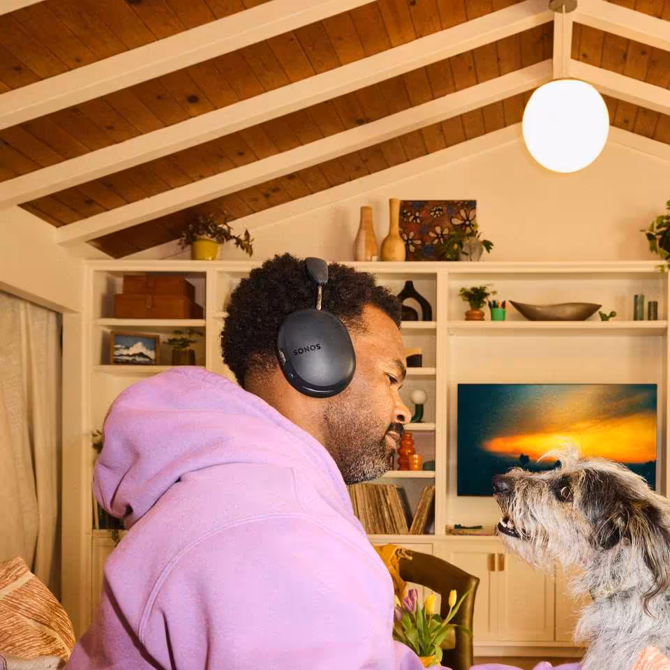 Personne portant un casque Sonos noir dans un salon, face à un chien