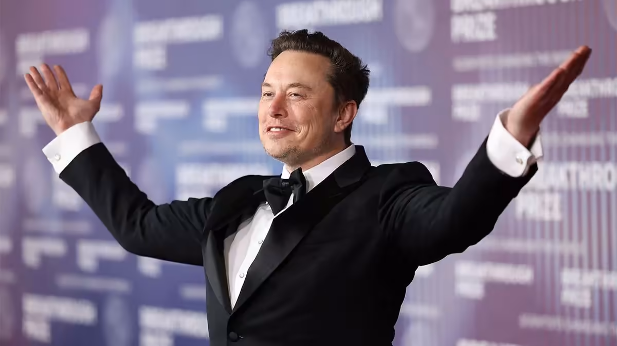 Terafab : Elon Musk reconnaît ne pas savoir fabriquer des semi-conducteurs 2 Elon Musk en costume noir, bras levés, devant un mur d’événement flou